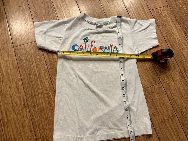 May include: White t-shirt with a colorful "California" graphic featuring palm trees, a surfboard, an orange, a flag, and a heart. The shirt is being measured with a tape measure.