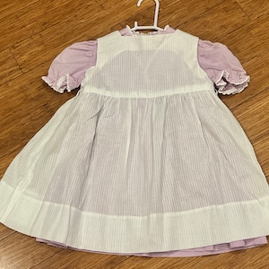 May include: A white and purple striped dress with a white pinafore overlay. The dress has a round neckline and short sleeves with lace trim.