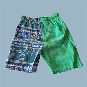 May include: Green and blue shorts with an elastic waistband. The left side features a blue and white patterned design with palm trees, a cartoon figure, and a volleyball. The right side is solid green. These shorts are perfect for casual wear.