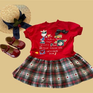May include: Red and plaid dress with short sleeves and a graphic print of a cartoon character on a school bus. The text on the dress reads "The school bus takes me for a ride, a good friend sits on either side!"
