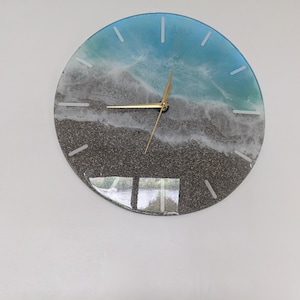 Puede incluir: Un reloj redondo con un diseño de escena de playa. La esfera del reloj está hecha de resina transparente con un diseño de ola azul y blanca y un fondo de arena gris. Las manecillas del reloj son doradas.
