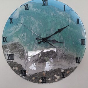 Peut inclure: Une horloge ronde avec des chiffres romains et des aiguilles noires. Le cadran de l'horloge est une coulée de résine avec un motif d'ondes océaniques bleues et blanches et du sable blanc et des coquillages en bas.