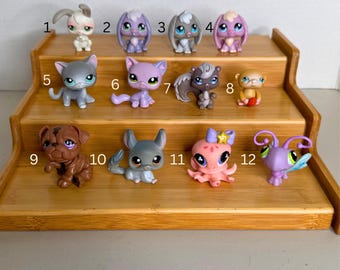 May include: A collection of twelve small plastic animal figurines arranged on a three-tiered wooden display stand. The figures include various creatures like rabbits, cats, a squirrel, a monkey, a dog, a chinchilla, an octopus, and a butterfly-like insect.