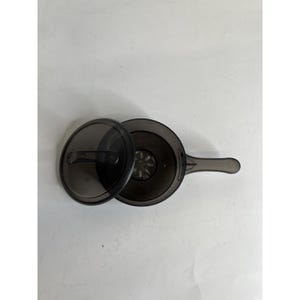 Op de afbeelding: Een doorschijnende donkergrijze plastic steelpan met bijpassend deksel. De pan heeft een lange steel en een ronde vorm. Het deksel is gedeeltelijk verwijderd, waardoor de ronde bodem met een gestructureerd, stervormig patroon zichtbaar is.