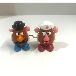 Può includere: Due giocattoli a carica Mr. Potato Head. Uno ha un cappello nero, baffi, pantaloni blu e naso arancione. L'altro ha un cappello bianco, labbra rosa e scarpe rosse. Entrambi hanno teste marroni a forma di patata e orecchie rosa.