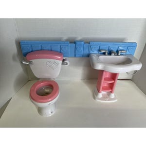 May include: A white and pink toy bathroom set featuring a toilet and a sink. The toilet has a pink seat and lid, and the sink has a pink pedestal. A blue wall piece with heart cutouts is above the sink and toilet.