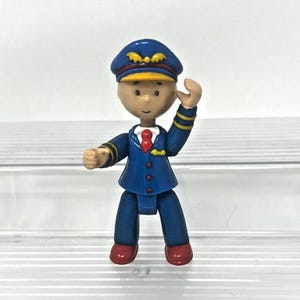 以下が含まれることがあります： 青い制服と帽子を身に着けたパイロットの小さなプラスチック製の置物。 パイロットは赤いネクタイと黄色のアクセントがあり、手を振っています。 置物は白い表面に立っています。