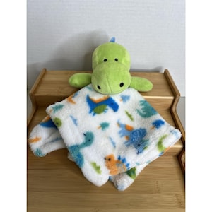 May include: A green dinosaur soft toy with a matching white and blue patterned blanket. The blanket has a soft, fluffy texture and is decorated with dinosaur illustrations in blue, orange, and green.