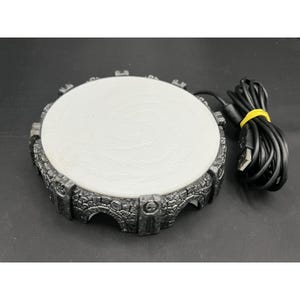 May include: A circular video game portal in grey and black, featuring a white central disc and a USB cable. The portal has a stone-like texture around the edge and is designed to connect to a gaming console.