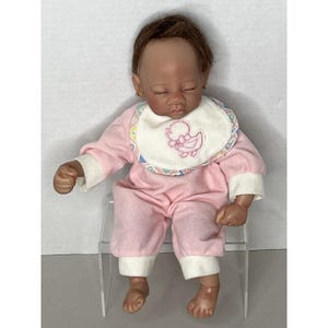 May include: A baby doll with brown hair and closed eyes, wearing a pink onesie with white cuffs and a bib featuring a duck design. The doll has small gold earrings and is seated on a clear plastic chair.