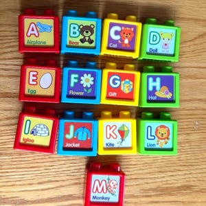 May include: A set of thirteen colourful plastic building blocks, each featuring a letter of the alphabet from A to M, accompanied by a corresponding illustration and word, such as Airplane, Bear, Cat, Doll, Egg, Flower, Gift, Hat, Igloo, Jacket, Kite, Lion, and Monkey.