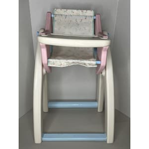 May include: A vintage doll high chair in pastel colors. The chair has a cream-colored frame with pink and light blue accents. The seat and backrest are covered in a floral patterned fabric. The chair is designed for small dolls.