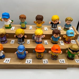 May include: A collection of fifteen small plastic toy figures, each depicting a person in various outfits and professions. The figures are arranged on a tiered wooden display, with each figure numbered for identification. The figures have different hair colors and wear hats.