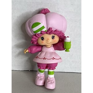 May include: A vintage Strawberry Shortcake figurine. The doll is wearing a pink dress, bloomers, and shoes, with green and white striped socks. She has a pink bonnet and holds a green milkshake with a cherry on top.