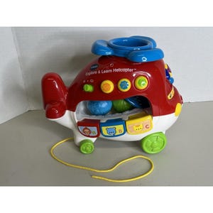 May include: A red, white, and blue plastic toy helicopter with the text "Explore & Learn Helicopter". The toy has a yellow pull string, green wheels, and colourful buttons. It features a blue rotor and a compartment with colourful balls.