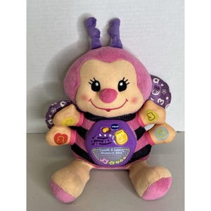 May include: A soft toy bee in pink, purple, and black. It has a smiling face, antennae, and wings with the number 3. The toy has a purple center with the text "Touch & Learn Musical Bee". It is designed for interactive play.