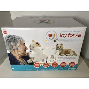 以下が含まれることがあります： Ageless Innovation Joy for Allコンパニオンペット用の白い長方形の段ボール箱。箱には高齢者とオレンジ色のタビーキャットのグラフィックが描かれており、Joy for Allというテキストと多言語の製品説明が記載されています。