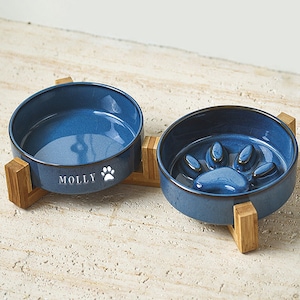 Może przedstawiać: Dwie niebieskie ceramiczne miski dla zwierząt na drewnianych stojakach. Jedna miska ma imię "MOLLY" i odcisk łapy. Druga miska ma wzór łapy na środku. Miski są w kolorze granatowym z brązową obwódką.