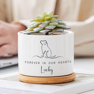 May include: White ceramic planter with a black line drawing of a dog and the text "Forever in our hearts Lucky". The planter has a wooden base and is filled with a green succulent plant.