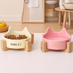 May include: Two cat-shaped pet bowls. One is cream-colored and filled with pet food, the other is pink. Both bowls are on wooden stands and have the name "Daisy" printed on them.