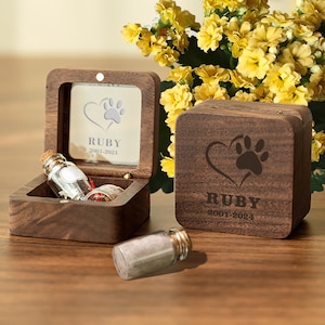May include: A wooden box with a heart and paw print design engraved on the lid. The box is personalized with the name "Ruby" and the dates "2001-2024". The box is open and contains two small glass vials.