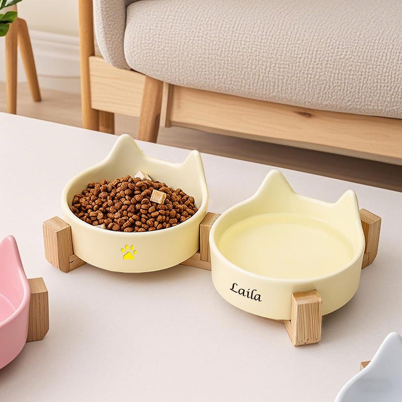 May include: Two cat-shaped ceramic pet bowls in a pale yellow color, one filled with dry pet food. The bowls sit on wooden stands. One bowl has a paw print, and the other has the name "Laila" printed on it.