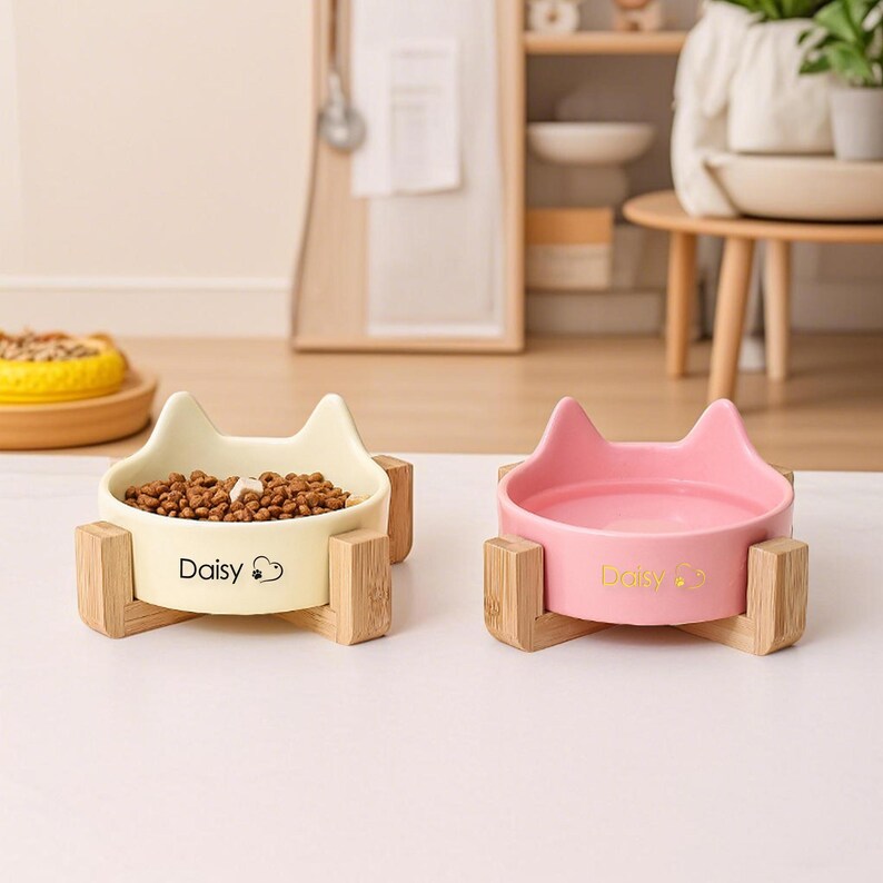 May include: Two cat-shaped pet bowls. One is cream-colored and filled with pet food, the other is pink. Both bowls are on wooden stands and have the name "Daisy" printed on them.