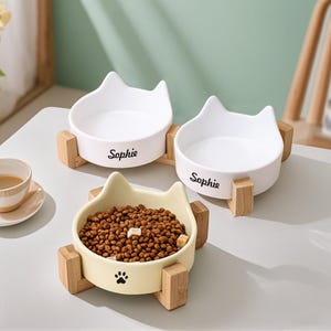 May include: Three ceramic cat-shaped pet bowls. Two white bowls with the name "Sophie" are empty, and one yellow bowl is filled with pet food and a paw print. Each bowl sits on a wooden stand.