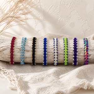 May include: Eight woven bracelets featuring intricate knot patterns and small beads. The bracelets are displayed in a row, showcasing various color combinations including deep red, light blue, black, royal blue, white, lime green, purple, and multicolored.