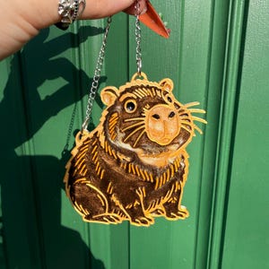 May include: A brown and gold capybara-shaped ornament suspended by a silver chain. The capybara features a detailed design, including whiskers and a smiling face. The ornament is set against a green backdrop.