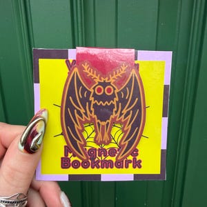 May include: A magnetic bookmark featuring a dark purple moth illustration with orange outlines and antennae, centered over a yellow spiderweb graphic. The backing card displays the text Magnetic Bookmark in purple letters against a yellow background.