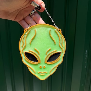 May include: A green alien head pendant with orange outlining and black eyes, suspended by a silver chain. The pendant has a smooth, glossy finish and is designed in a cartoon style. The background is a solid, dark green.