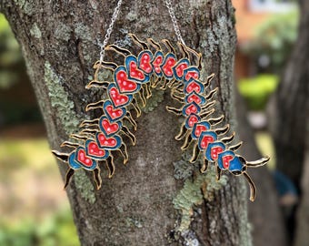 May include: A decorative hanging ornament shaped like a centipede with a segmented body. Each segment features a red heart with white dots, outlined in blue and black metal. The piece is suspended by a thin silver-colored metal chain.