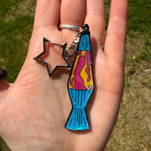 May include: A keychain featuring a lava lamp charm with a blue base and top, containing yellow and pink blobs. The charm has a holographic finish and is attached to a silver-colored star-shaped clasp by a small metal jump ring and lobster claw.