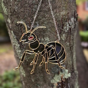May include: A hanging ornament shaped like an ant, crafted with black enamel and gold-toned metal outlines. The ant features a prominent red eye and is suspended by a thin silver-colored metal chain, showcasing a detailed insect silhouette.