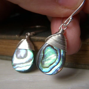 May include: A pair of silver wire wrapped earrings with iridescent abalone shell drops. The earrings have a teardrop shape and are a beautiful shade of blue and green.