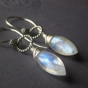 May include: A pair of silver earrings with teardrop-shaped, iridescent moonstone pendants. The earrings feature a twisted silver design and are suspended from a simple hook. The moonstones have a milky, opalescent quality.