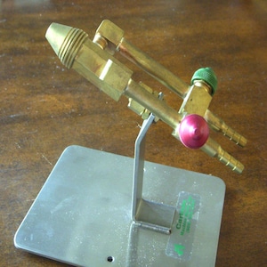 May include: A brass torch head with a red and green valve, mounted on a silver metal stand. The torch head has a ribbed nozzle and two tubes extending from the body. The stand has a rectangular base.