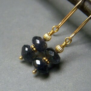 May include: Close-up of gold-colored earrings with dark blue, faceted beads. The earrings have a simple design with a hook and small gold accents. The beads are oval-shaped and have a polished, reflective surface. The jewelry is set against a gray background.