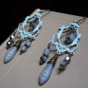 May include: A pair of light blue dangle earrings with a filigree design and iridescent blue beads. The earrings have a teardrop shape with a swirl pattern.