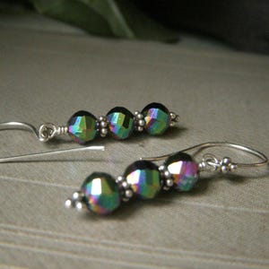 May include: A pair of dangle earrings featuring iridescent, faceted beads in shades of green, purple, and blue. The beads are strung on silver-colored wire, with small silver beads accenting the design. The earrings have a simple hook closure.