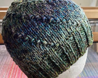 May include: A hand-knitted beanie hat in shades of green, blue, and purple. The hat features a textured, ribbed design and is displayed on a white mannequin head. The yarn has a variegated color pattern, creating a unique look.