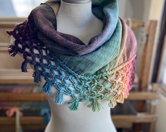 May include: A rainbow colored crocheted scarf with a knotted fringe. The scarf is displayed on a white mannequin.