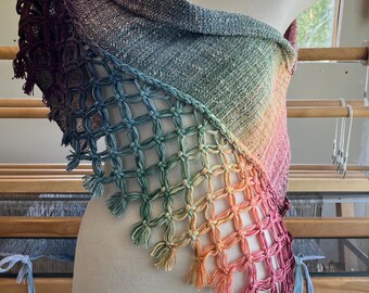 May include: A colorful shawl displayed on a mannequin. The shawl features a gradient of colors, transitioning from dark burgundy to light pink, with a woven net design along the edge and tassels. The background includes a window and a weaving loom.