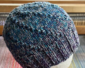 May include: A hand-knitted beanie hat in shades of blue, teal, purple, and white. The hat features a ribbed knit pattern and is displayed on a white mannequin head. The yarn appears to be a blend of colors, creating a textured effect.