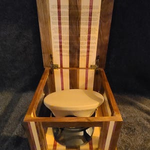 May include: Wooden box with a hinged lid, featuring alternating stripes of dark and light wood with purple and white accents. Inside, a cream-colored hat rests on a silver stand. The lid's interior displays printed text.