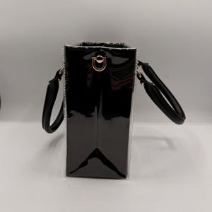 May include: A structured black handbag with a glossy patent finish and clear side panels. The bag features two rounded black handles and gold-toned metal hardware, including a D-ring attachment point on the side for a shoulder strap.