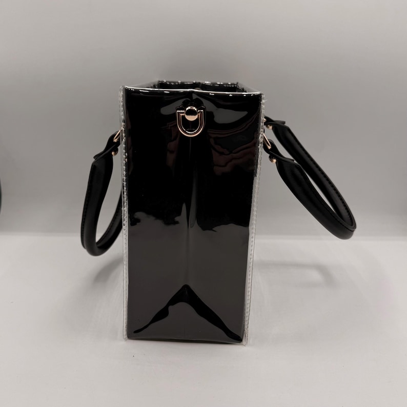 May include: A structured black handbag with a glossy patent finish and clear side panels. The bag features two rounded black handles and gold-toned metal hardware, including a D-ring attachment point on the side for a shoulder strap.