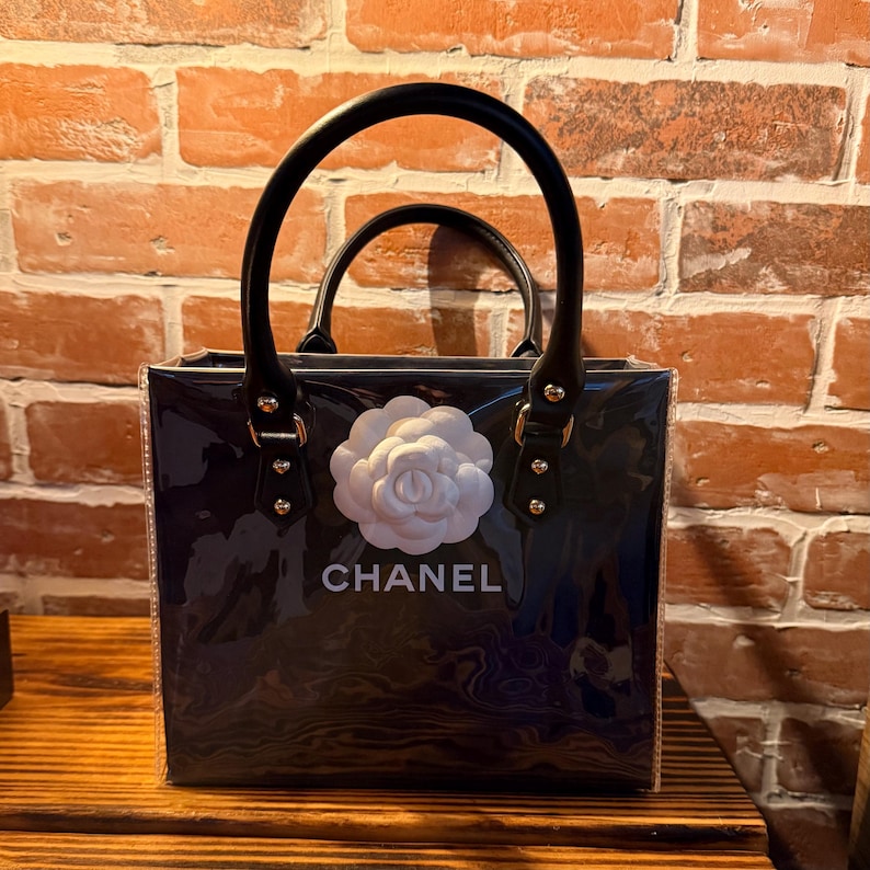 May include: A square-shaped, transparent dark-toned handbag featuring two black rounded handles attached with gold-tone hardware. A white three-dimensional camellia flower is centered on the front, positioned above the white text CHANEL.