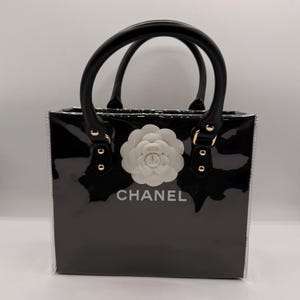 May include: A black glossy handbag with a structured rectangular shape, featuring two black top handles with gold-tone hardware. A large white camellia flower emblem is centered on the front, positioned above the white text CHANEL.
